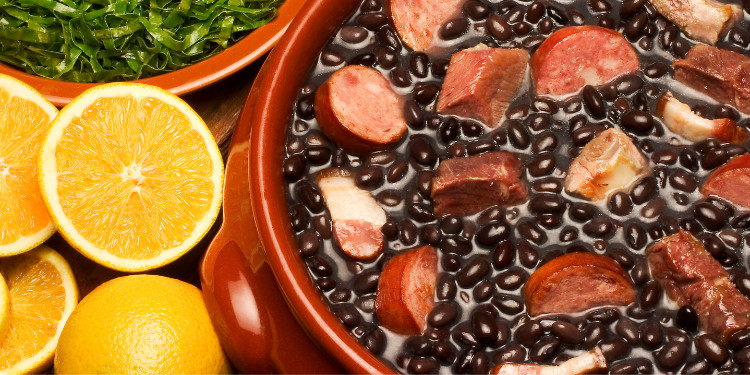 Feijoada brasileira em panela de barro com laranja cortada ao lado e couve em tigela.