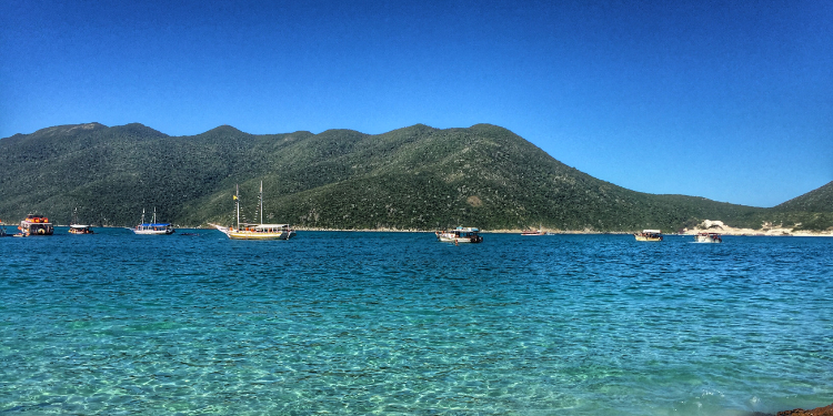 Fotografia da orla de Arraial do Cabo, mostrando água azul turquesa transparente, montanhas verdejantes ao fundo e diversos barcos ancorados na baía sob um céu azul claro.