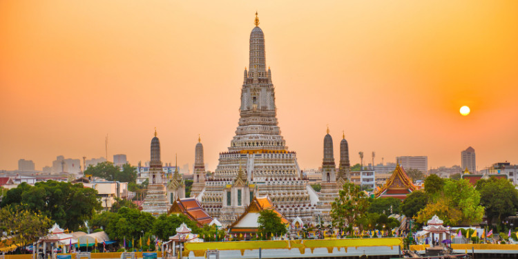 Fotografia do templo Wat Arun em Bangkok, Tailândia, ao pôr do sol, com a silhueta do templo contra um céu laranja vibrante, o sol poente no canto superior direito e o rio Chao Phraya em primeiro plano.