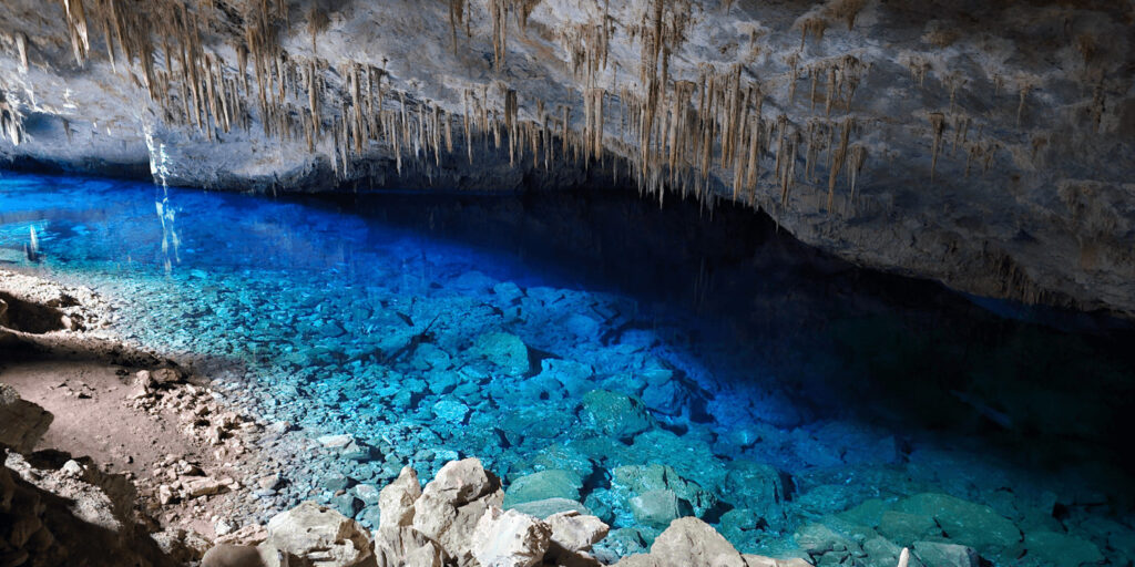 ma caverna com um lago azul cristalino em Bonito, MS. Estalactites pendem do teto da caverna e rochas são visíveis nas águas rasas.