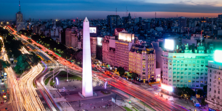 Fotografia noturna do Obelisco de Buenos Aires, Argentina, iluminado em branco, com rastros de luz de carros na Avenida 9 de Julio e edifícios da cidade ao fundo sob um céu noturno azulado.