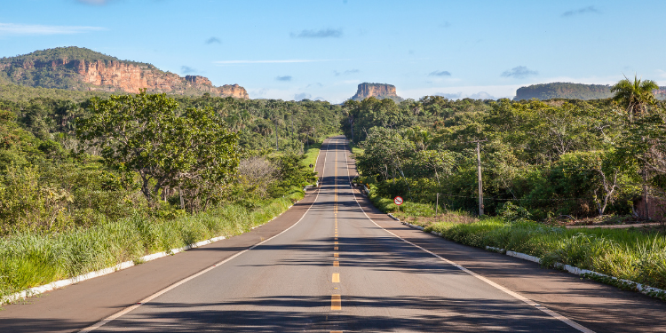 Fotografia de uma estrada de asfalto reta cortando uma paisagem verdejante com montanhas de topo plano (mesas) ao fundo, característica da Chapada das Mesas.