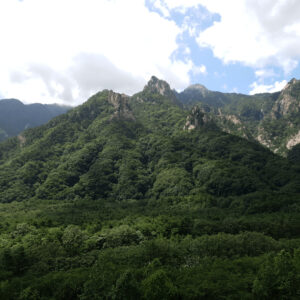 Montanhas com formações rochosas distintas e densa vegetação, com nuvens no céu azul, criando um cenário dramático.