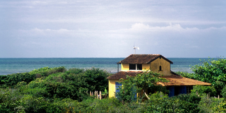 Fotografia de uma casa amarela com telhado marrom, cercada por vegetação verde, com o oceano azul claro ao fundo, representando a arquitetura típica e a beleza natural da Ilha do Cardoso.
