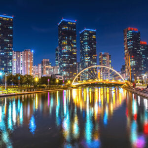 Vista noturna do Central Park de Songdo, com prédios modernos iluminados ao fundo e o lago refletindo as luzes da cidade.