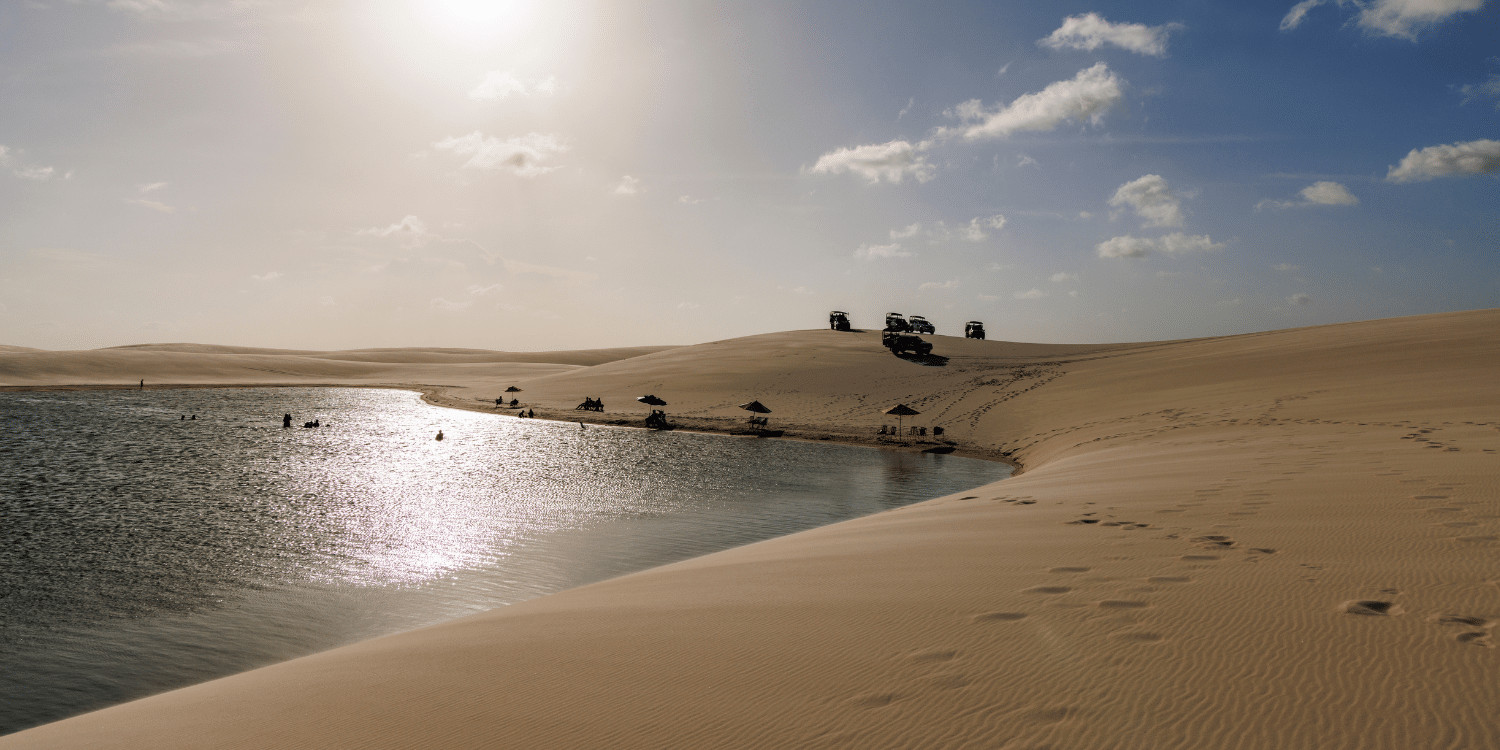 Fotografia de paisagem dos Lençóis Maranhenses, Brasil, com uma lagoa de água azul refletindo o céu, cercada por dunas de areia branca, e veículos 4x4 e pessoas na duna ao fundo.