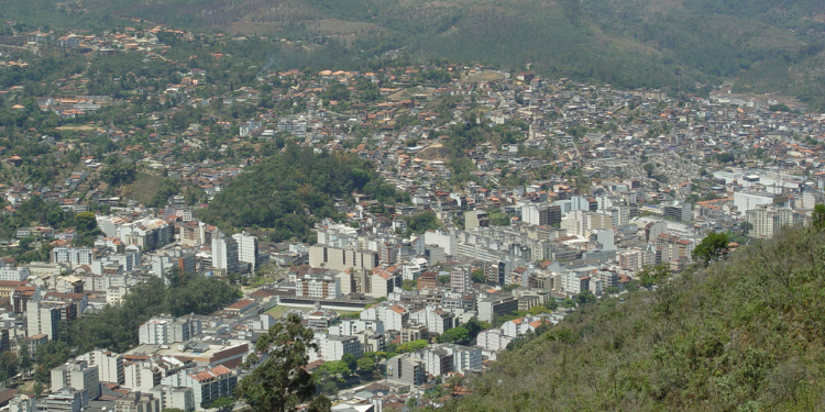 Fotografia aérea de Nova Friburgo, RJ, com edifícios e casas densamente construídos em um vale cercado por montanhas cobertas de vegetação verde, sob um céu nublado.