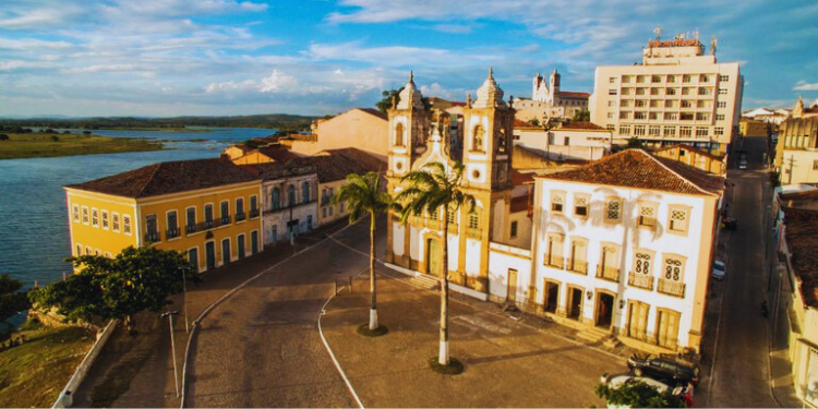 Fotografia aérea de Penedo, Alagoas, mostrando a Igreja Matriz com suas torres gêmeas brancas e amarelas, cercada por edifícios coloniais coloridos e o Rio São Francisco ao fundo.