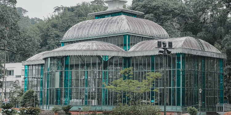 Fotografia do Palácio de Cristal em Petrópolis, RJ, mostrando a estrutura de vidro e metal com detalhes ornamentados, cúpulas e claraboias, cercado por árvores verdes e vegetação densa, sob um céu nublado.