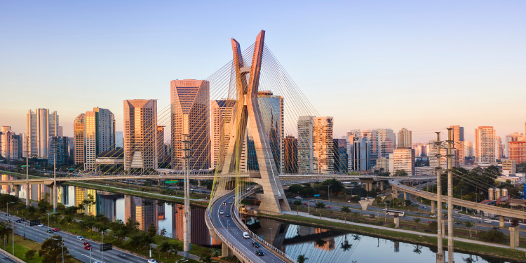 Fotografia aérea da Ponte Estaiada Octávio Frias de Oliveira em São Paulo, com seus cabos tensionados e pilares em "X" destacados, edifícios altos em segundo plano, vias expressas e o rio Pinheiros visíveis, tudo sob um céu claro com tons de rosa do amanhecer.