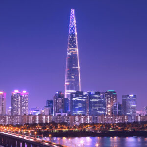 Uma foto noturna do Lotte World Tower, um arranha-céu altíssimo com design moderno e iluminação vibrante, destacando-se contra o céu noturno de Seul, com outras luzes da cidade visíveis em segundo plano.