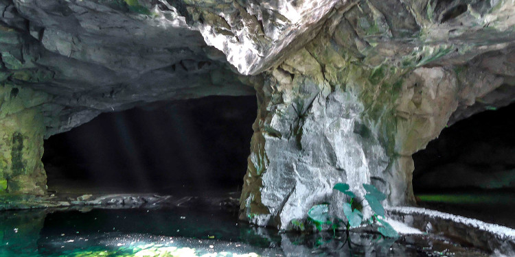 Interior de uma gruta com lago de água verde-esmeralda, formações rochosas e luzes que penetram a caverna.
