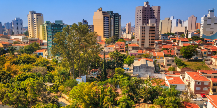 Fotografia aérea de Sorocaba, SP, exibindo prédios altos de diferentes cores e alturas, casas com telhados de cerâmica laranja, árvores verdes e vegetação densa, tudo sob um céu azul claro com poucas nuvens.