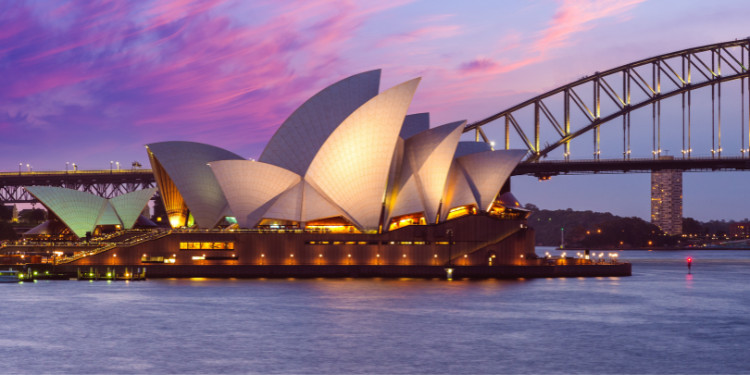 Ópera de Sydney, Austrália, iluminada à noite com suas velas brancas distintas. A Harbour Bridge, com luzes douradas, está ao fundo e o céu apresenta tons de roxo e rosa.