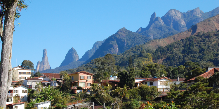 Fotografia de Teresópolis, RJ, mostrando casas de tons claros com telhados de cerâmica, árvores verdejantes e a Serra dos Órgãos com seus picos rochosos e íngremes ao fundo, sob um céu azul claro.