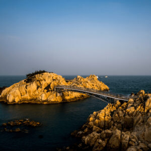 Uma vista aérea da ponte pênsil Daewangam, uma estrutura moderna de metal que se estende por uma enseada rochosa, com o oceano e o céu azul ao fundo.