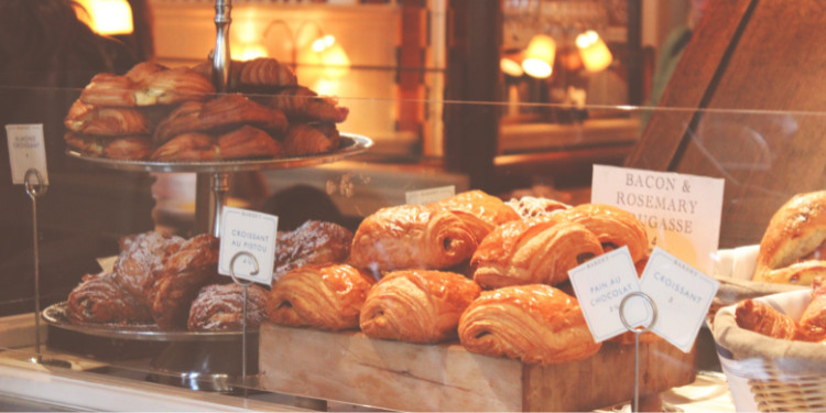 itrine de padaria com croissants e outros pães em pratos e cestas, com placa 'BACON & ROSEMARY MIGASSE'.