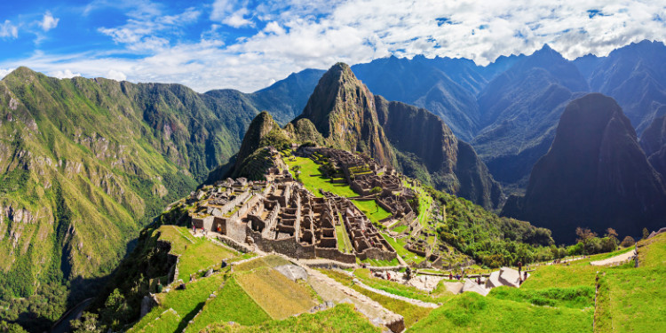 Vista panorâmica das ruínas de Machu Picchu, Peru, com estruturas de pedra em terraços, Wayna Picchu ao fundo, montanhas verdejantes e céu azul com nuvens brancas.