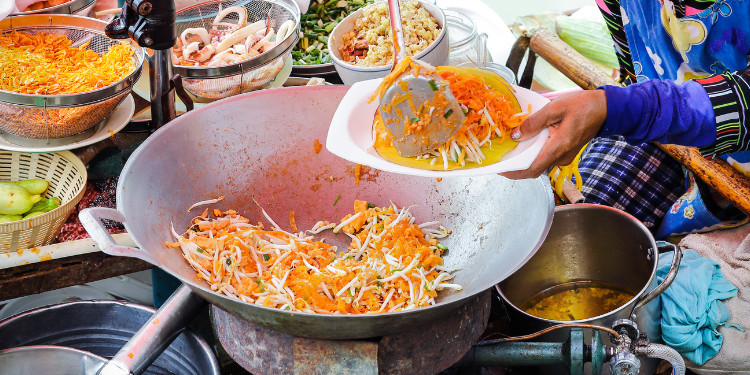 Uma pessoa servindo Pad Thai em um prato, com uma grande wok de metal e outros ingredientes visíveis ao fundo.