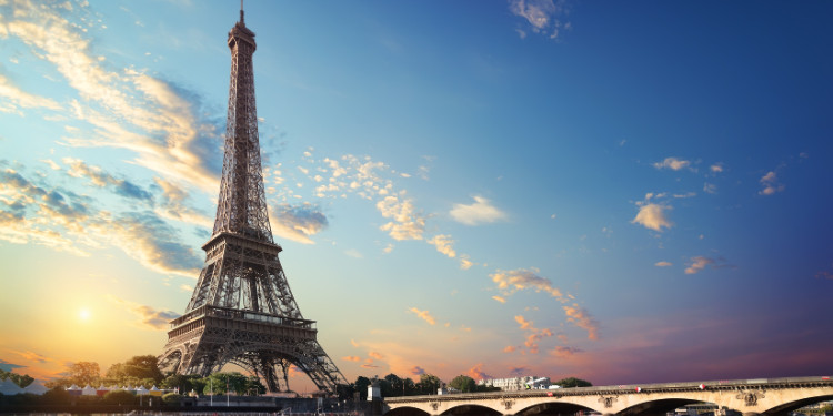 Fotografia da Torre Eiffel em Paris, França, com o sol se pondo atrás dela, criando um céu com tons de laranja e rosa. A torre se destaca contra o céu e o Rio Sena pode ser visto em primeiro plano.