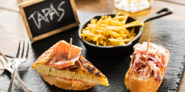 Tapas espanholas com tortilla, presunto e batatas fritas em prato de ardósia, com placa 'TAPAS' ao fundo.