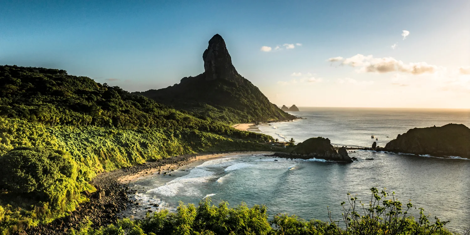 Vista panorâmica de Fernando de Noronha, com vegetação exuberante, praia de águas cristalinas e o icônico Morro do Pico ao fundo.