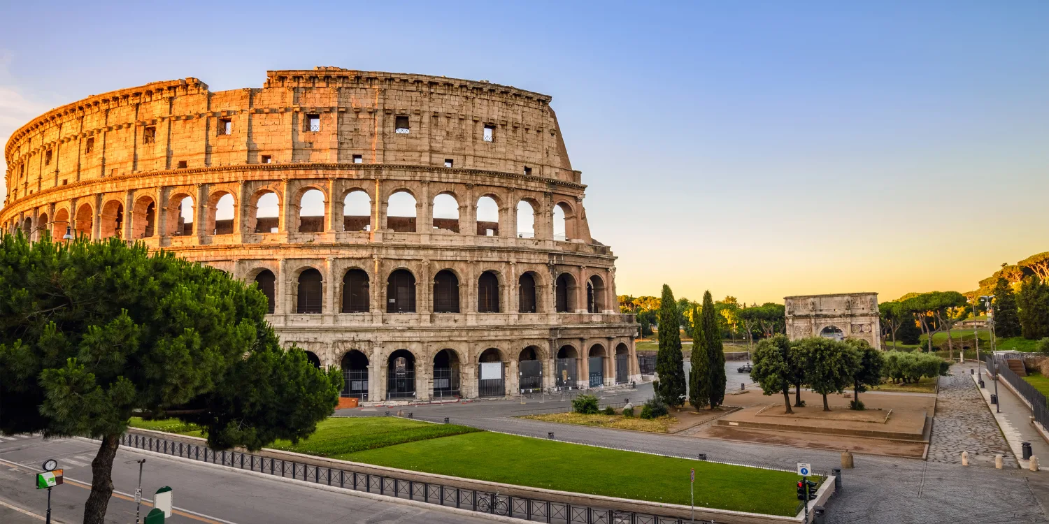 Coliseu de Roma iluminado pela luz do pôr do sol, com céu azul e poucas pessoas ao redor.