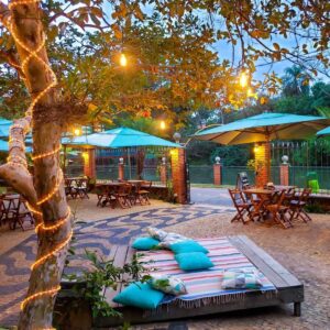 Foto do Kanoas Bar em Brotas, mostrando um espaço ao ar livre com mesas de madeira, guarda-sóis, luzes aconchegantes e um deck com almofadas coloridas. A atmosfera é convidativa e relaxante, perfeita para desfrutar de momentos especiais.