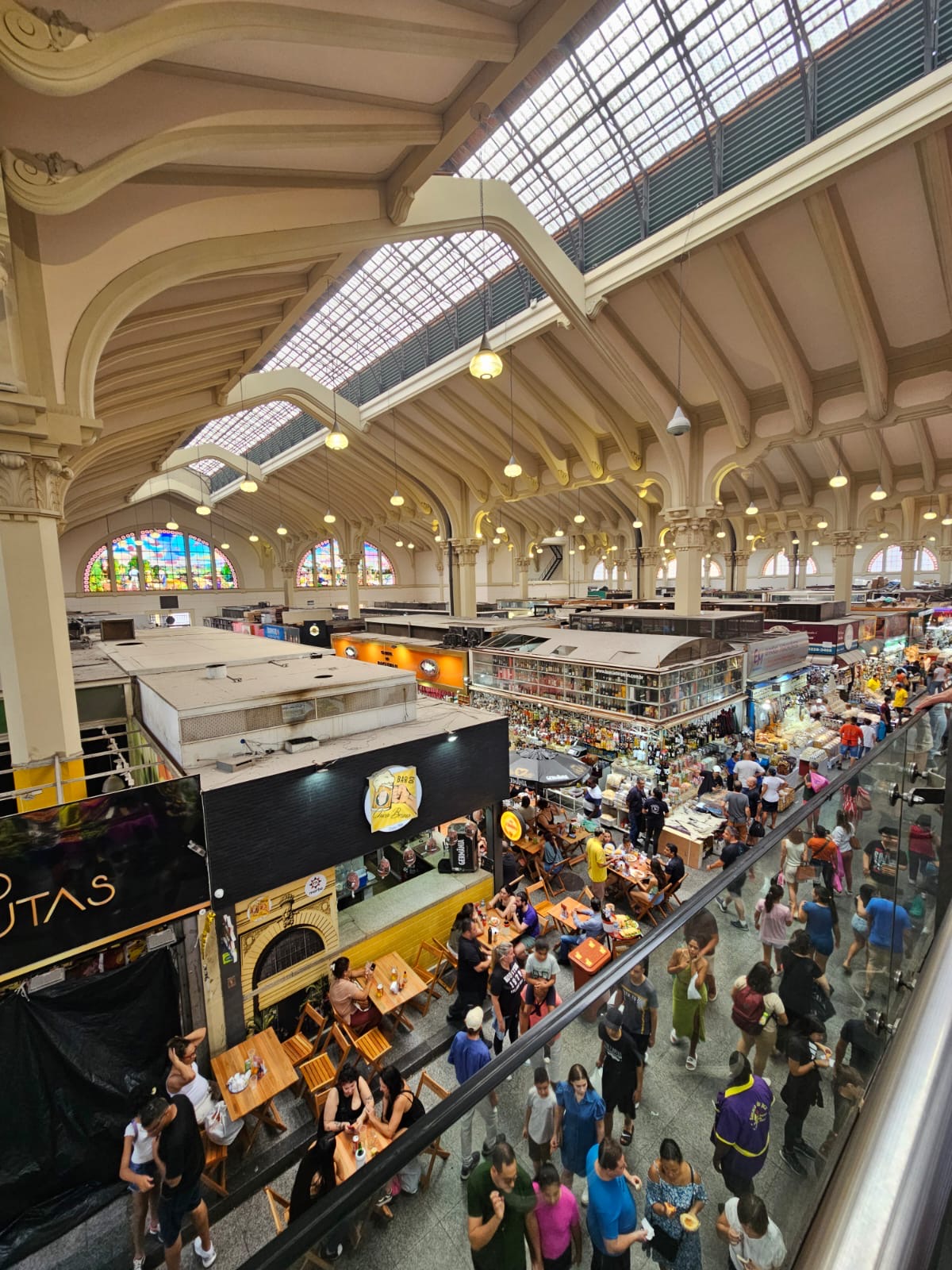 Fotografia do interior do Mercado Municipal de São Paulo, mostrando a arquitetura detalhada do teto com seus arcos e claraboias, bancas de frutas e alimentos, e pessoas caminhando e sentadas.