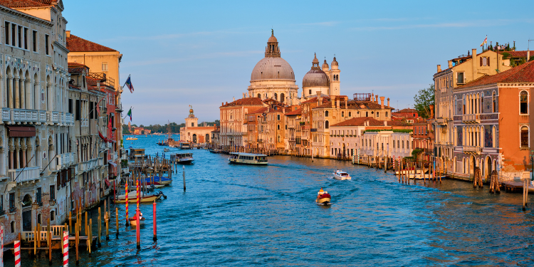 Vista do Grande Canal em Veneza, mostrando edifícios históricos ao longo das margens, com a Basílica de Santa Maria della Salute ao fundo, barcos e gôndolas navegando nas águas, sob um céu claro ao entardecer.