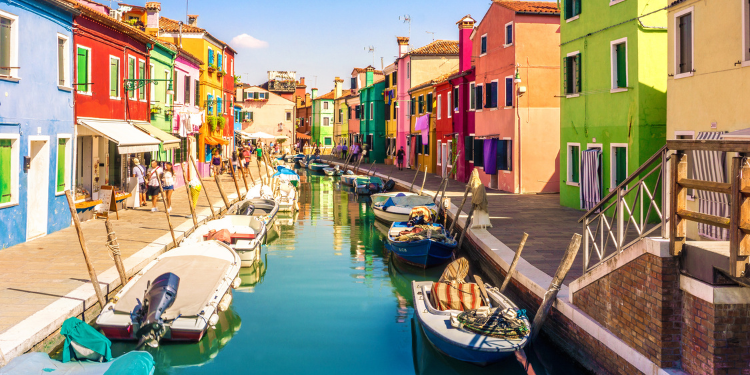 Rua colorida em Burano, Veneza, com casas vibrantes pintadas em diversas cores, um canal estreito com barcos estacionados ao centro, e pessoas caminhando ao longo das margens.