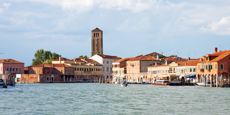 Paisagem urbana de Murano, Veneza, com edifícios de tijolos laranja e vermelhos ao longo de um canal de água, uma torre sineira alta ao fundo, e barcos atracados ao longo da margem.