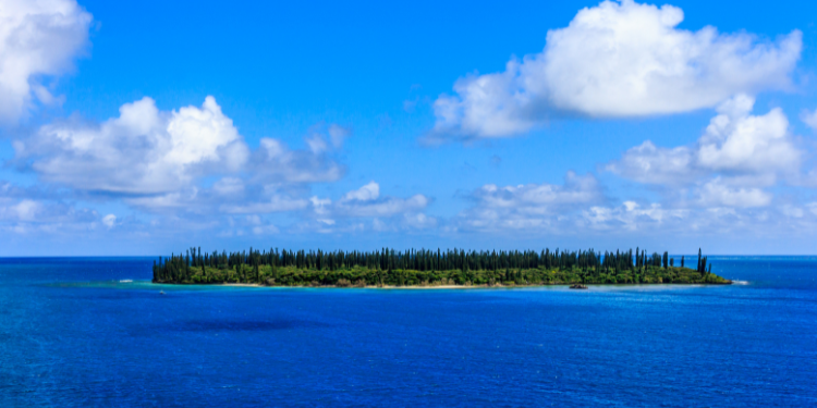 ista panorâmica da Ilha dos Pinheiros, Nova Caledônia, com uma pequena ilha coberta de pinheiros em meio a um mar azul profundo, sob um céu claro com nuvens brancas.