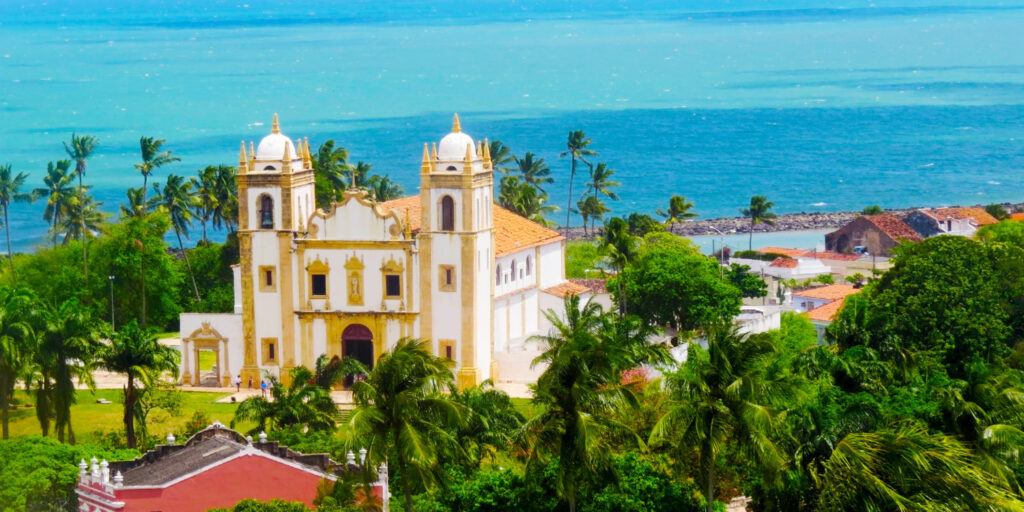 Fotografia aérea da Igreja do Carmo em Olinda, Brasil, com suas torres brancas e detalhes amarelos, cercada por palmeiras e vegetação verde, com o mar azul turquesa ao fundo.