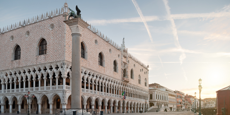 Fachada do Palácio Ducal em Veneza, mostrando detalhes arquitetônicos góticos, incluindo arcos e colunas, com uma escultura de leão no topo de uma coluna, sob um céu claro com rastros de avião.