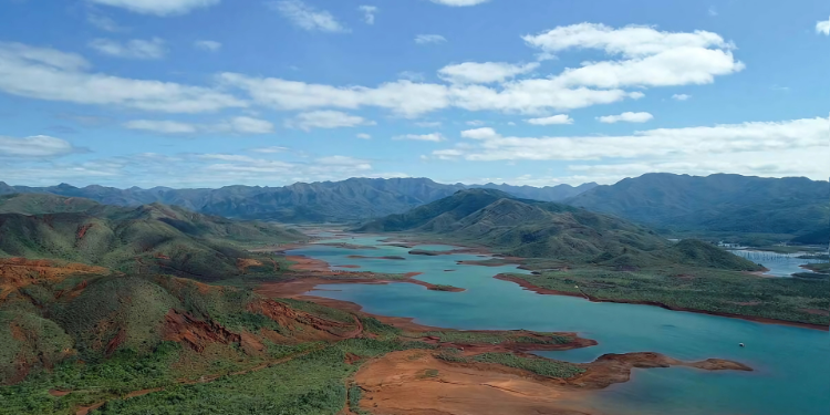 Visão aérea do Parque Provincial da Rivière Bleue, mostrando um rio de águas turquesas serpenteando por entre montanhas cobertas de vegetação verde e áreas de terra vermelha, sob um céu azul com nuvens brancas.