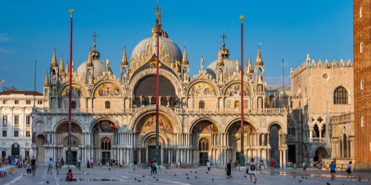 Vista da Piazza San Marco em Veneza, mostrando a Basílica de São Marcos com suas cúpulas e detalhes arquitetônicos ornamentados, o campanário ao lado, e pessoas caminhando na praça sob um céu claro.