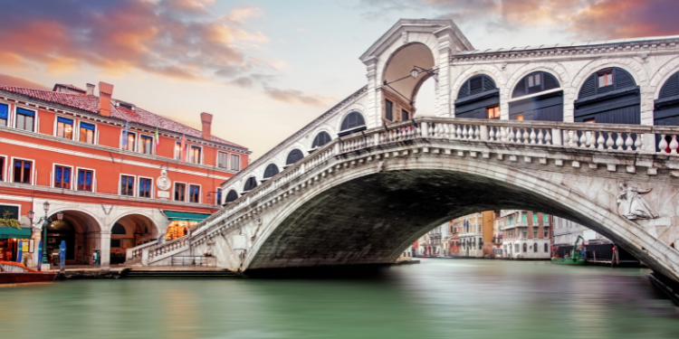 Ponte Rialto em Veneza, mostrando sua arquitetura em arco sobre o Grande Canal, com edifícios coloridos ao fundo e o reflexo da ponte na água.