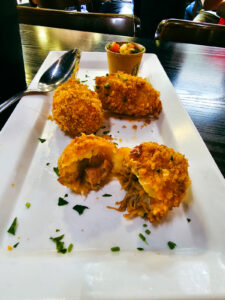 Fotografia de quatro bolinhos de costela fritos, com casquinha dourada e crocante, recheados com carne desfiada, um deles cortado ao meio para mostrar o recheio, acompanhados de vinagrete em um pequeno pote e salsa picada espalhada no prato.