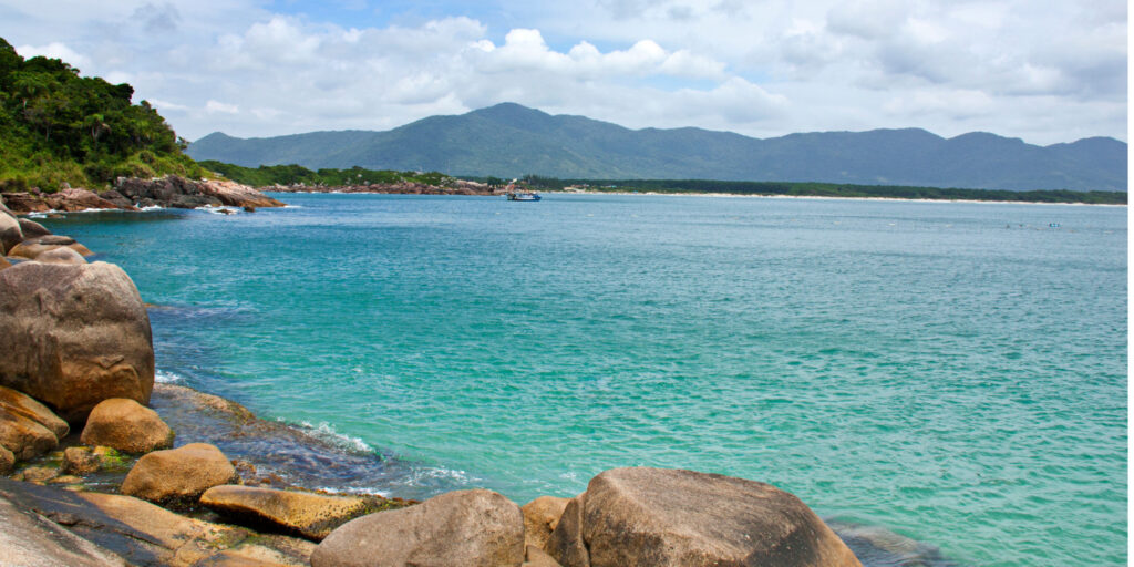 Fotografia de Florianópolis, Brasil, mostrando uma baía com água azul turquesa clara, grandes pedras na costa e montanhas verdes ao fundo sob um céu parcialmente nublado.