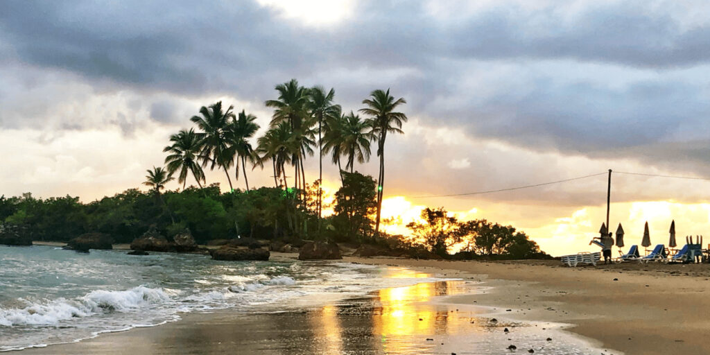 Fotografia de uma praia em Morro de São Paulo, Brasil, ao entardecer, com palmeiras altas no centro, ondas suaves na areia molhada, e um céu nublado com um brilho alaranjado do sol poente refletido na areia molhada.