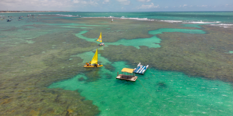Vista aérea das piscinas naturais de Porto de Galinhas, mostrando recifes de coral na maré baixa, águas claras com tons de azul e verde, e pequenas embarcações coloridas navegando.