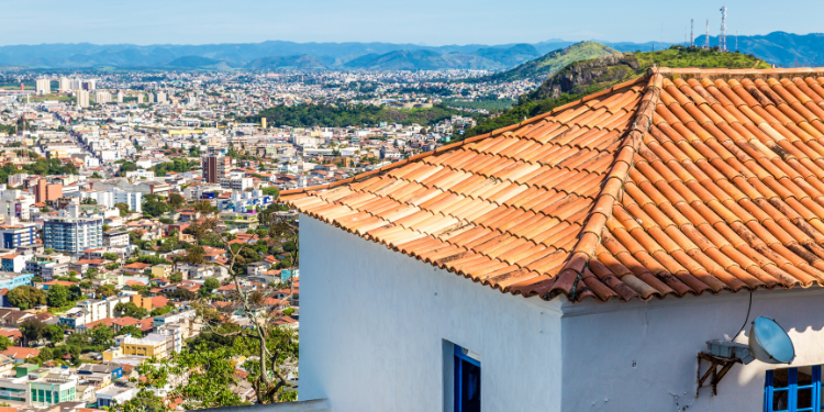 Uma vista aérea parcial da cidade de Vitória, Espírito Santo, com o telhado de uma casa colonial de telhas de barro em primeiro plano, e o panorama urbano da cidade com morros e montanhas ao fundo.