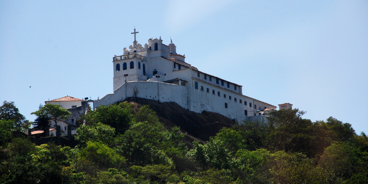 O Convento da Penha, uma construção branca imponente com uma cruz no topo, situado no alto de um morro verdejante, contra um céu azul claro.
