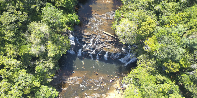 Vista aérea da Cachoeira do Tijuípe, na Bahia, mostrando a queda d'água em meio à Mata Atlântica, com diversas pessoas nadando no rio e no poço principal.